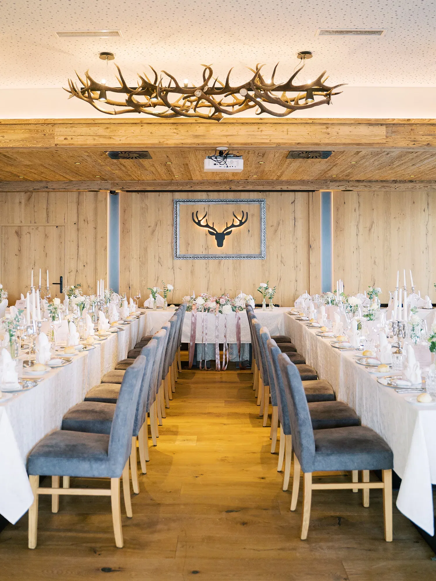 Hochzeitssaal im Landhausstil mit langen Tafeln, geschmückt mit Blumen und Kerzen, der Raum wird von einer Holzdecke und einem Hirschgeweih dominiert.