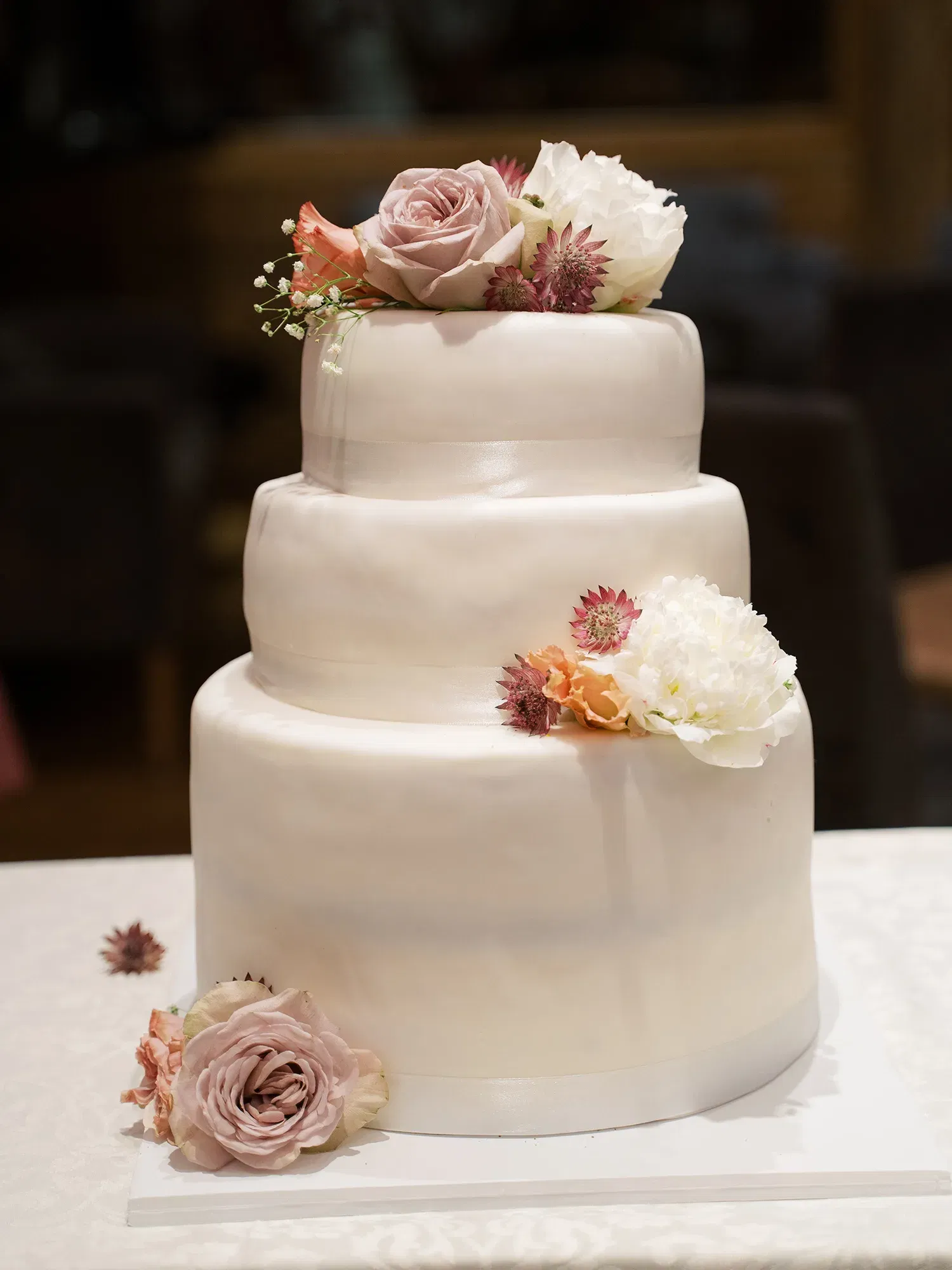 Schlichte, dreistöckige Hochzeitstorte mit Blumen in zarten Rosa- und Weißtönen, dekorativ arrangiert auf einem mit Blumenblättern geschmückten Tisch.