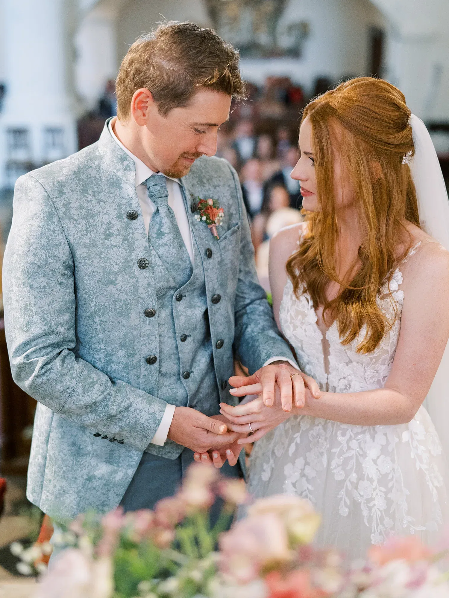 Hochzeitspaar während des Ringtauschs, der Bräutigam steckt der Braut in einem Moment der Zärtlichkeit den Ehering an, umgeben von Hochzeitsgästen.