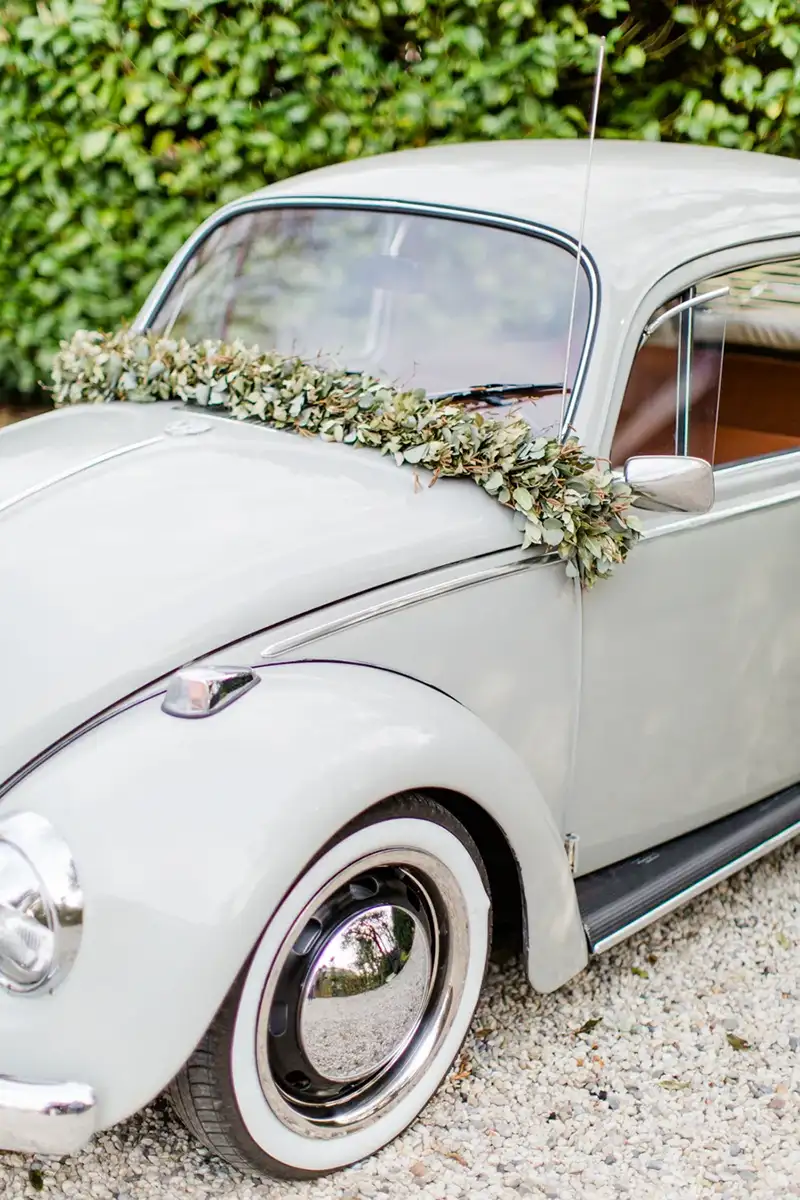 Ein stilvoller VW Käfer, dekoriert mit einem floralen Kranz, steht auf einer Kiesauffahrt für den Hochzeitstag bereit.