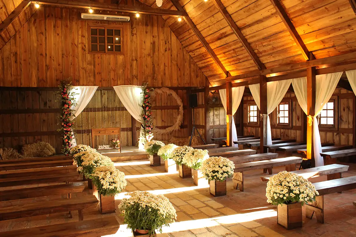 Ein rustikaler Holzsaal, der für eine Feier oder Hochzeit dekoriert ist. Der Raum hat eine warme Atmosphäre mit Holzbalken an der Decke, Blumendekorationen entlang des Ganges und Holzbänken, die zum Altar führen. Die Lichtstrahlen tauchen den Raum in sanftes, goldenes Licht.