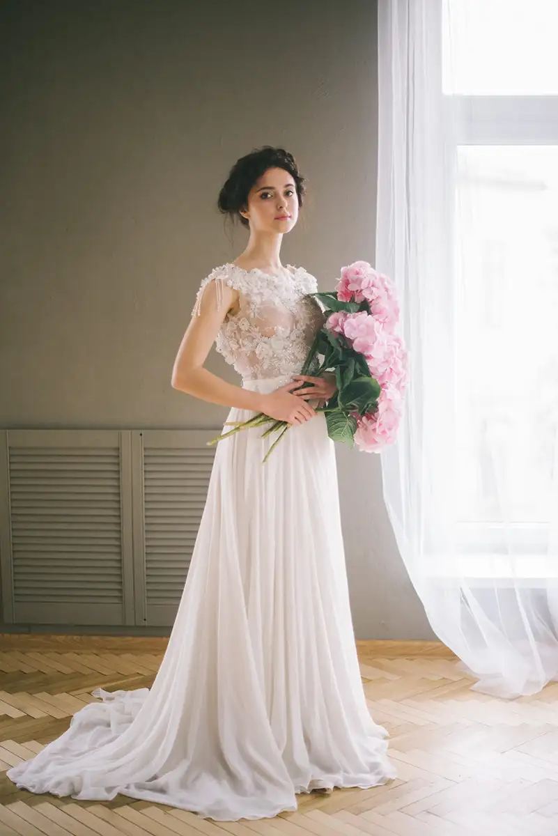 Eine Braut in einem bodenlangen, weißen Kleid hält einen üppigen Strauß mit rosa Pfingstrosen. Sie posiert vor einem lichtdurchfluteten Fenster.