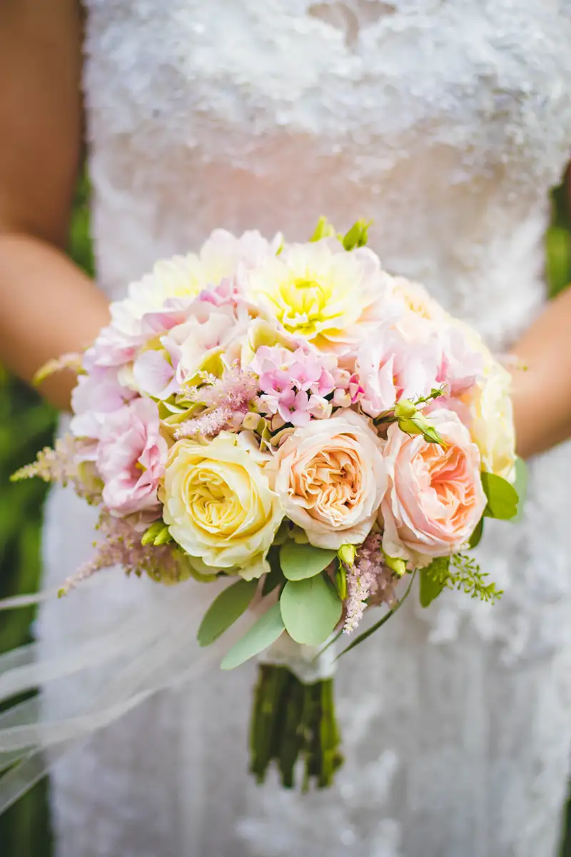 Romantisches Hochzeitsbouquet mit rosa und cremefarbenen Rosen, kombiniert mit zarten grünen Blättern, getragen von einer Braut in einem weißen Kleid.