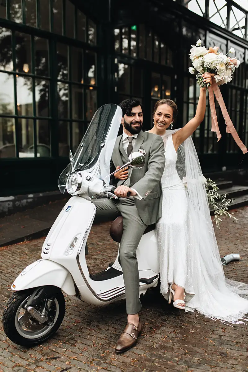 Ein Brautpaar sitzt auf einem weißen Roller und strahlt vor Freude, die Braut schwenkt ihren Brautstrauß, während sie sich auf den gemeinsamen Weg macht.