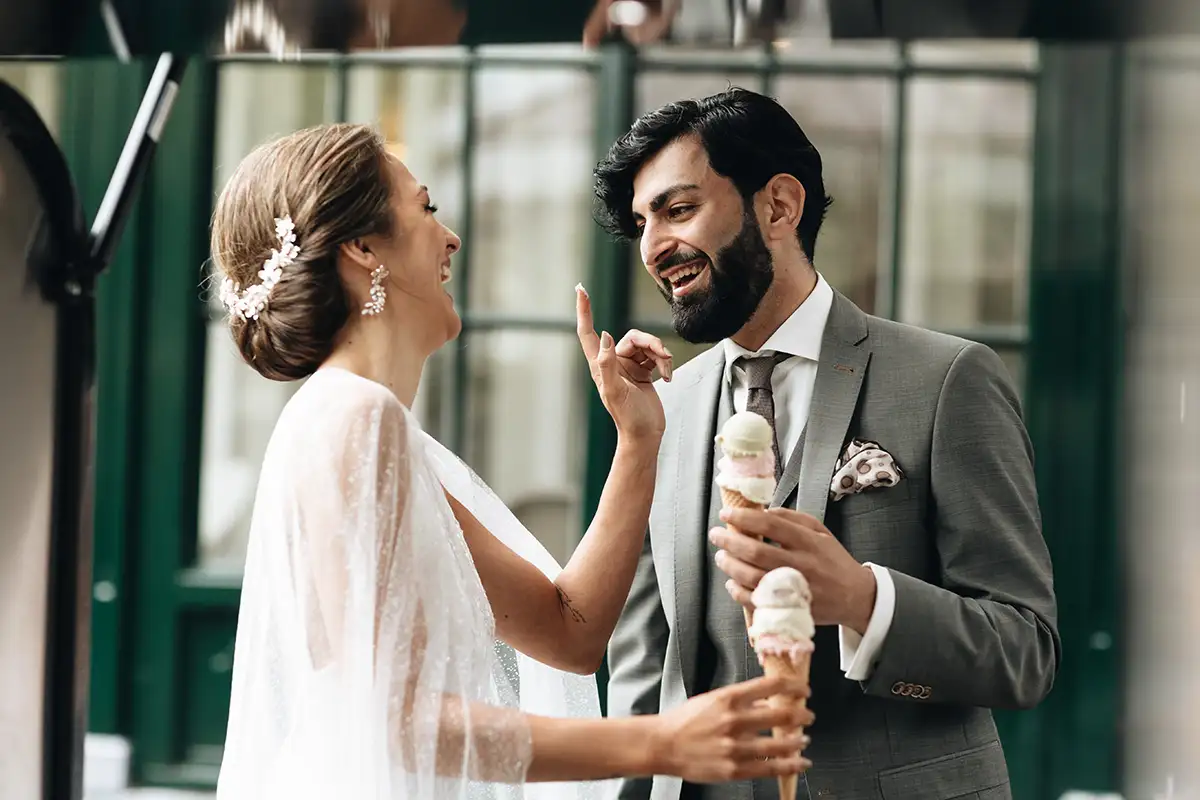 Hochzeitspaar, das fröhlich Eis isst, die Braut neckt den Bräutigam spielerisch, während sie beide elegante Hochzeitskleidung tragen.