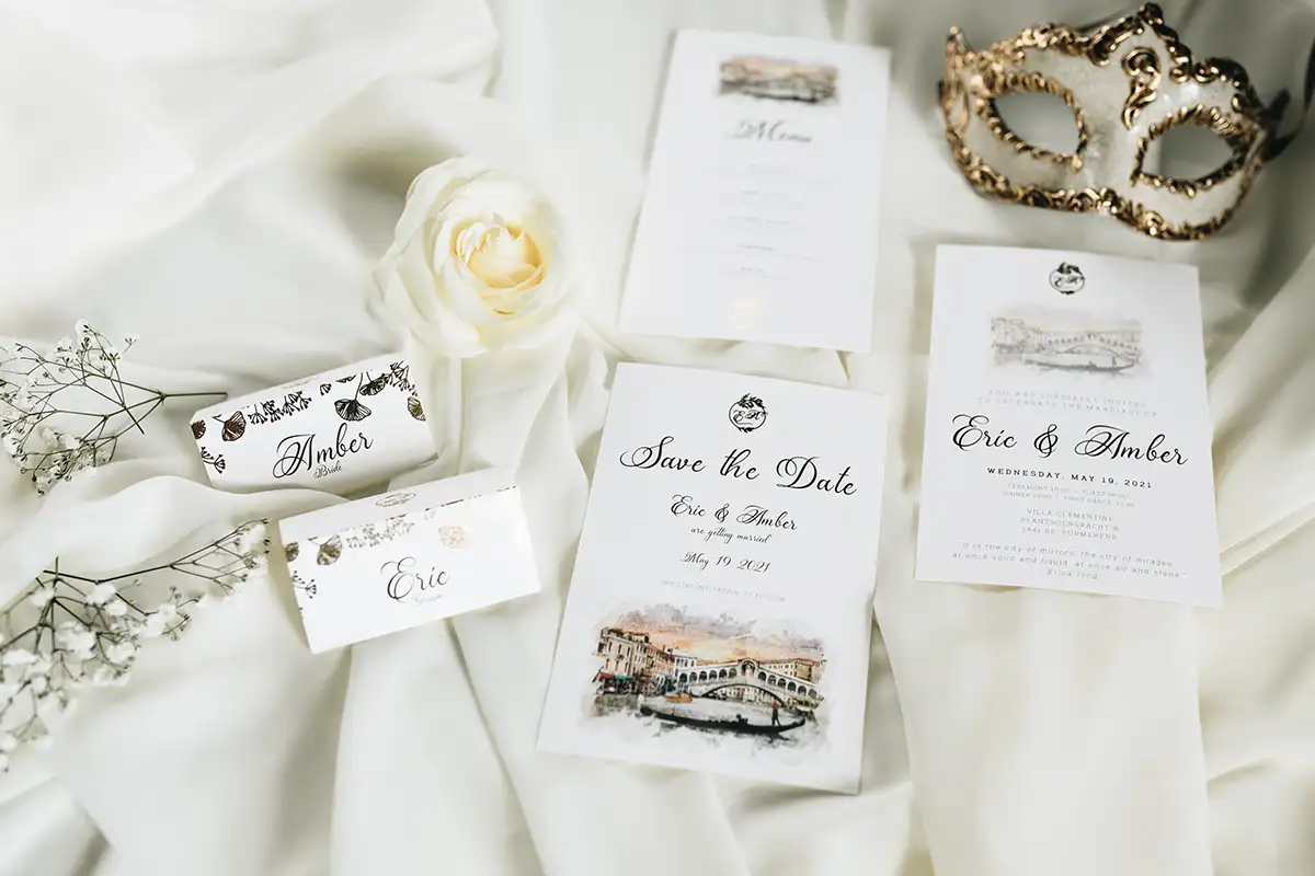 Einladungskarten und Tischkarten-Set, arrangiert auf einem weißen Tuch, umgeben von einer weißen Rose und einer dekorativen Maske, die dem Thema der Hochzeit Eleganz verleihen.