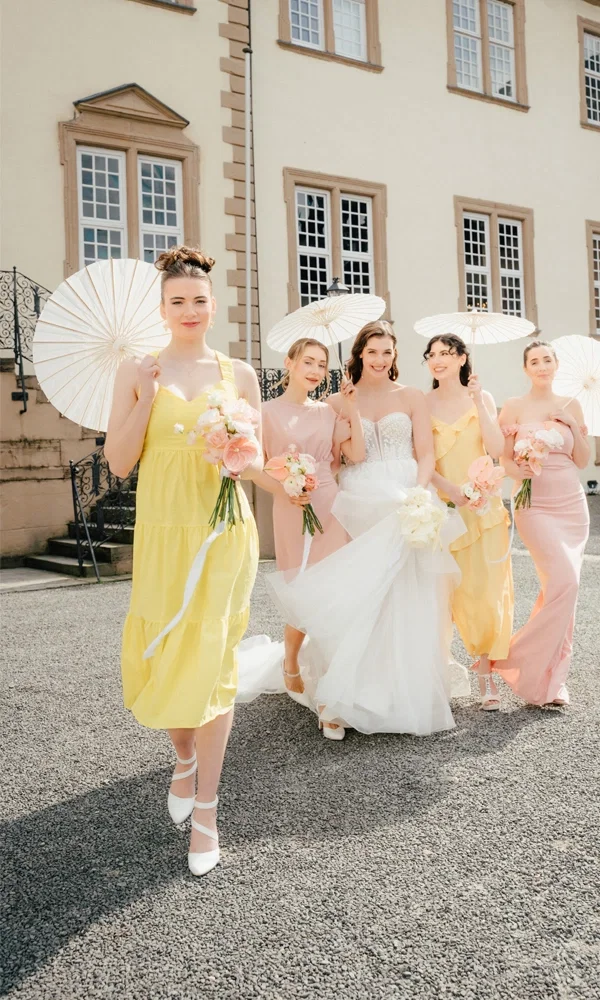 Eine fröhliche Gruppe bestehend aus einer Braut und ihren Brautjungfern posiert vor einer prächtigen Villa. Die Braut trägt ein schulterfreies weißes Kleid, während die Brautjungfern farbenfrohe Sommerkleider in Gelb und Rosa tragen. Alle halten Blumensträuße und Sonnenschirme, die zu der entspannten, sommerlichen Stimmung der Hochzeitsfeier passen.