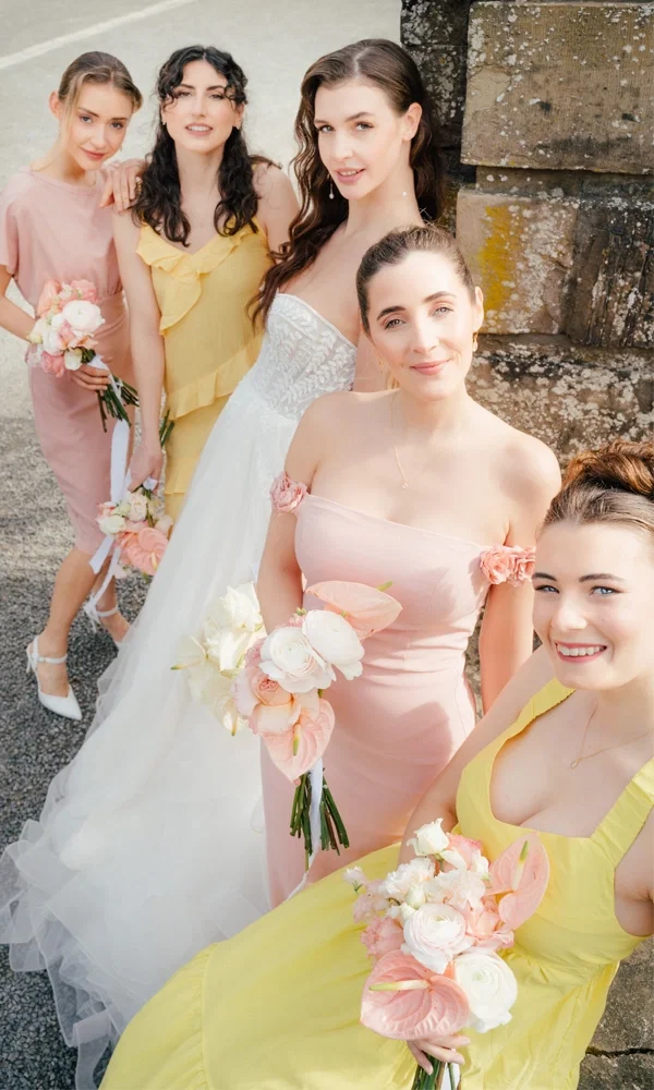 Eine Braut in einem trägerlosen weißen Kleid posiert mit ihren vier Brautjungfern, die helle, sommerliche Kleider in Gelb und Rosa tragen. Alle halten Blumensträuße aus pastellfarbenen Blüten. Die Gruppe steht in enger Formation vor einem alten, steinernen Hintergrund, die Stimmung wirkt freundlich und entspannt, passend zur sommerlichen Hochzeitsfeier.