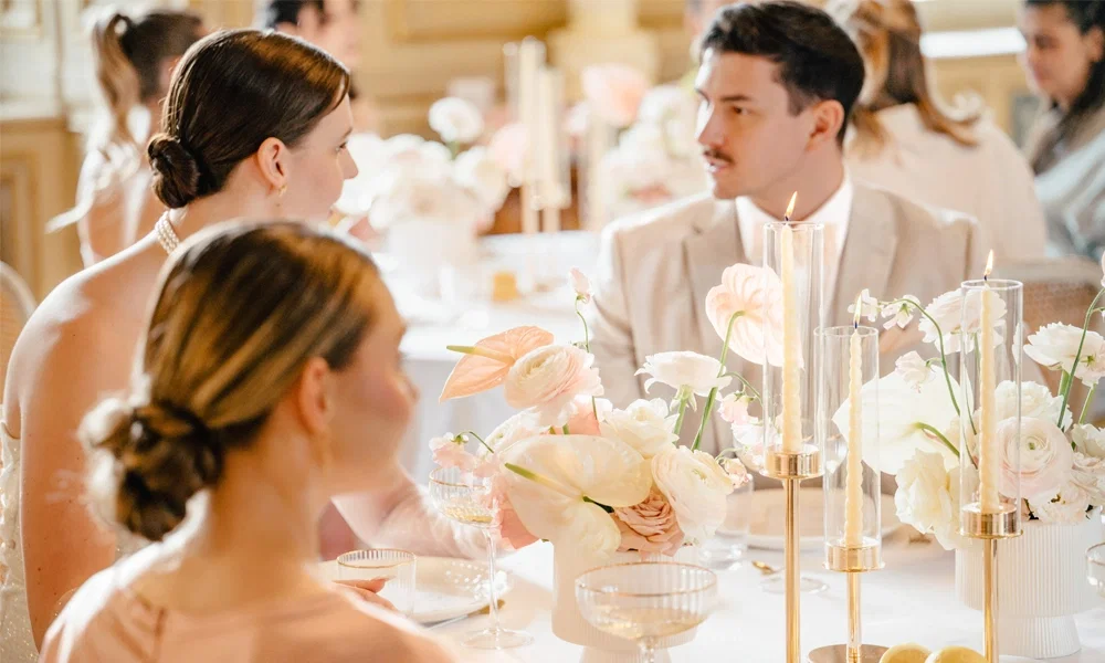 Eine feierliche Szene mit mehreren Personen, die sich an einem edel gedeckten Tisch unterhalten. Die Tischmitte ist mit blassen Blumenarrangements und stilvollen Kerzenleuchtern geschmückt. Auf dem Tisch liegen Zitronenhälften, und es sind elegante Weingläser zu sehen, während warme Kerzenlichter eine gemütliche Atmosphäre erzeugen.