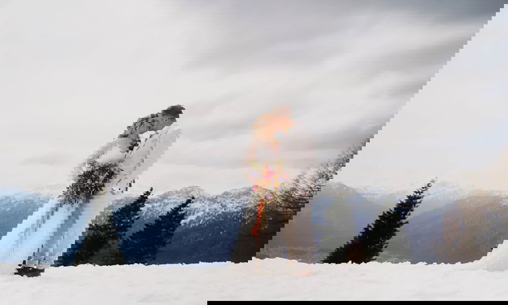 Ein frisch verheiratetes Paar steht im Schnee vor einer atemberaubenden Bergkulisse. Die Braut mit langem Spitzenkleid und Blumenkranz hält einen lebhaften Brautstrauß mit bunten Blüten. Der Bräutigam, in einem beigen Anzug, beugt sich sanft zu ihr hinunter für einen innigen Kuss.