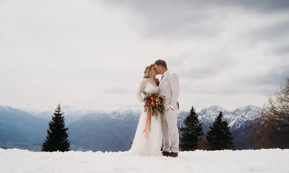 Ein frisch verheiratetes Paar steht im Schnee vor einer atemberaubenden Bergkulisse. Die Braut mit langem Spitzenkleid und Blumenkranz hält einen lebhaften Brautstrauß mit bunten Blüten. Der Bräutigam, in einem beigen Anzug, beugt sich sanft zu ihr hinunter für einen innigen Kuss.