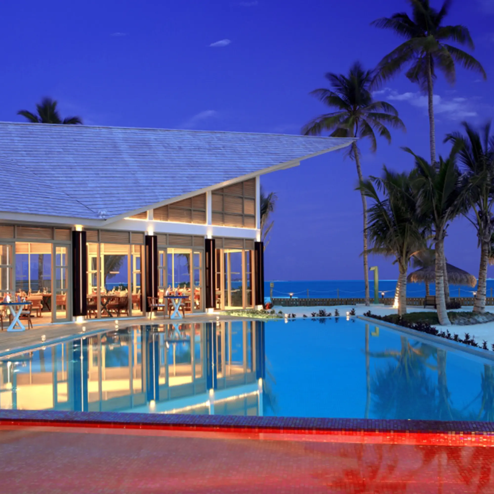 Romantisches Abendessen am Strand bei Nacht, mit beleuchteten Tischen unter freiem Himmel und dem sanften Rauschen der Wellen im Hintergrund.