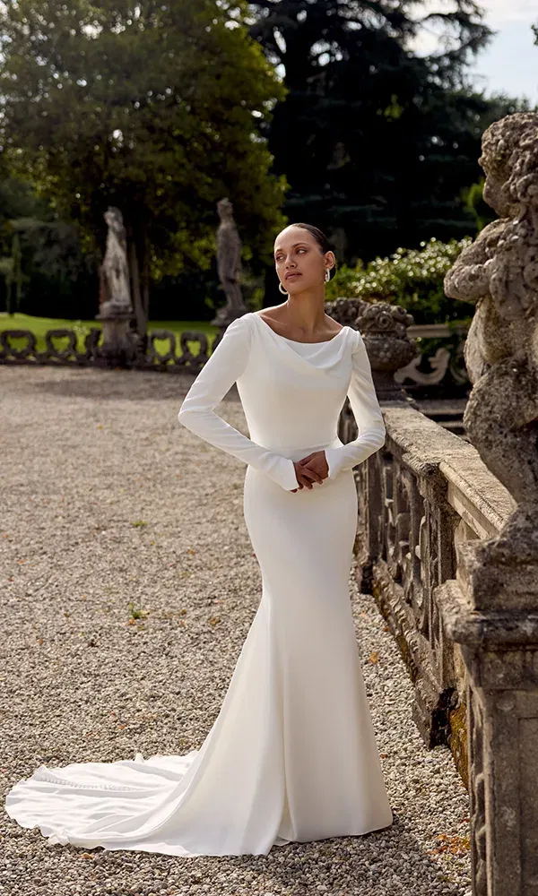 Zeitlose Eleganz: Figurbetontes Brautkleid mit langen Ärmeln und U-Boot-Ausschnitt, getragen im Schlossgarten mit Statuen im Hintergrund.