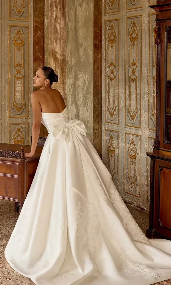 Prunkvolles Brautkleid mit opulenter Schleppe und XXL-Schleife am Rücken, inszeniert in einem prachtvollen, goldverzierten Raum.