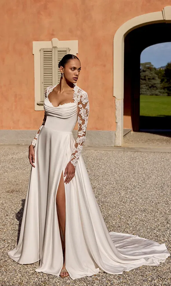 Modernes Brautkleid mit Schlitz und langen, transparenten Spitzenärmeln, aufgenommen im Innenhof vor einer toskanischen Villa.