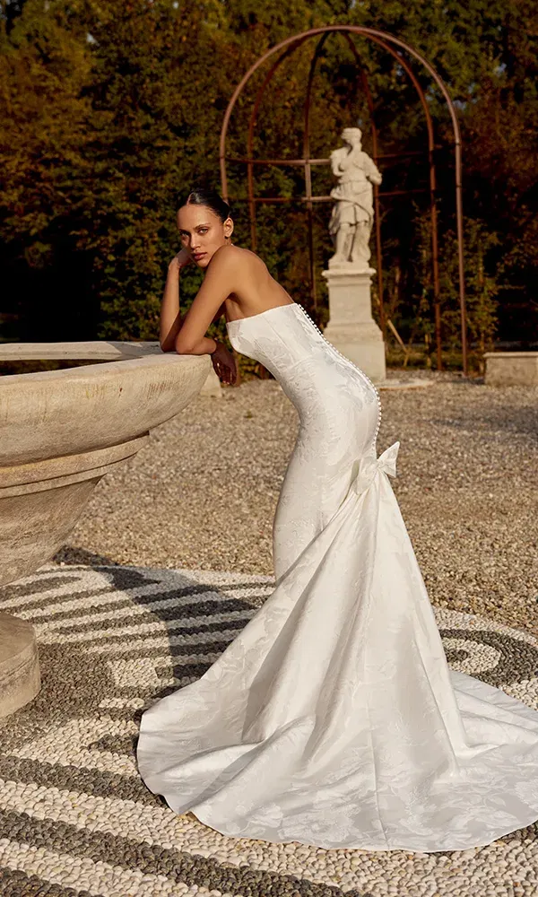 Figurbetontes Jacquard-Kleid mit Schleife und Schleppe, stilvoll in Szene gesetzt an einem steinernen Brunnen im Schlossgarten.