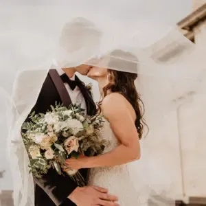 Brautpaar küsst sich unter einem weißen Schleier und die Braut hält zeitgleich einen massiven Brautstrauß.