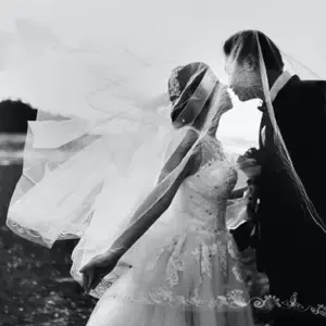 Die Braut küsst unter einem transparenten Schleier den Bräutigam in schwraz-weiß.