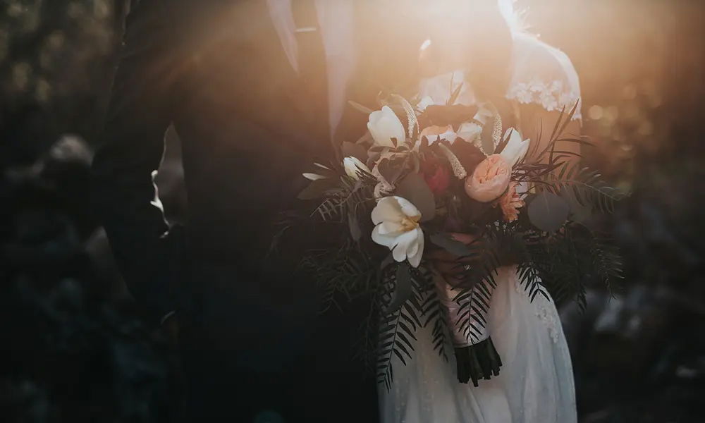 Ein romantischer Moment im Gegenlicht: Die Braut präsentiert einen großen Blumenstrauß mit weißen Lilien, Rosen und grünen Akzenten. Das Brautpaar wird von warmem Sonnenlicht sanft umspielt, das für eine traumhafte Atmosphäre sorgt.