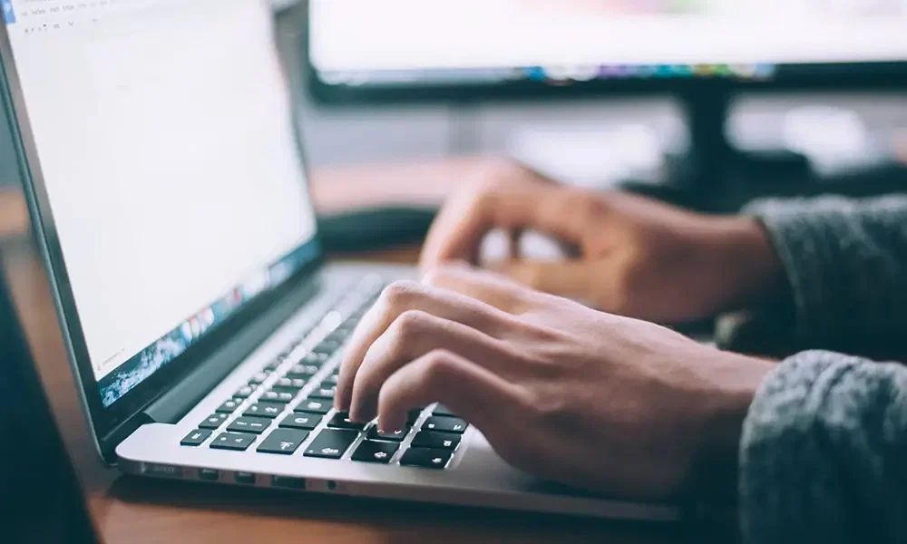Eine Person arbeitet konzentriert an einem Laptop: Die Hände tippen auf der Tastatur, während im Hintergrund ein Computerbildschirm sichtbar ist.