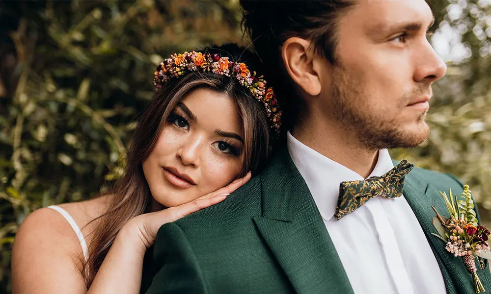 Nahaufnahme eines Brautpaares: Die Braut mit zarten Blumen im Haar blickt liebevoll in die Kamera, während der Bräutigam mit ernstem Ausdruck in die Ferne schaut.