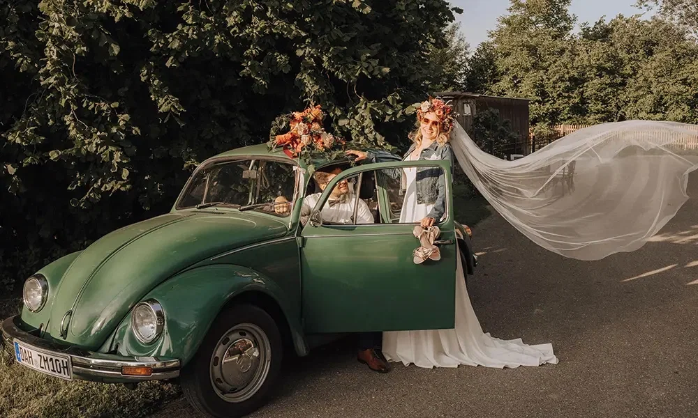 Hochzeitsfoto mit Retro-Charme: Eine Braut steht neben einem klassischen grünen VW Käfer, ihr Schleier fliegt dramatisch in die Luft. Der Bräutigam sitzt fröhlich am Steuer.