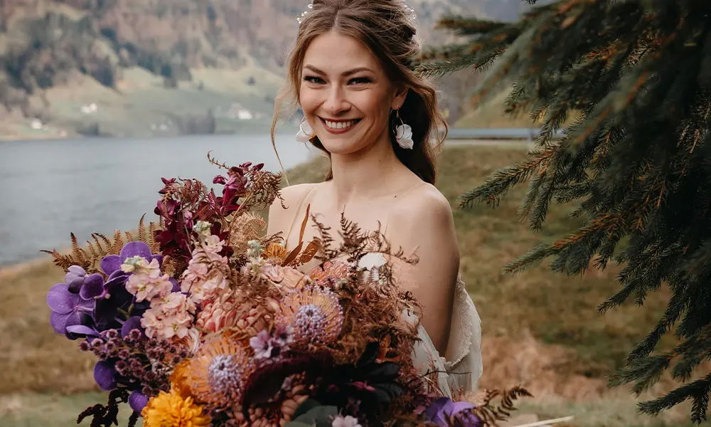 Eine glückliche Braut mit einem farbenfrohen, wilden Blumenstrauß posiert vor einer idyllischen Landschaft aus See und Bergen, eingerahmt von Tannenzweigen.