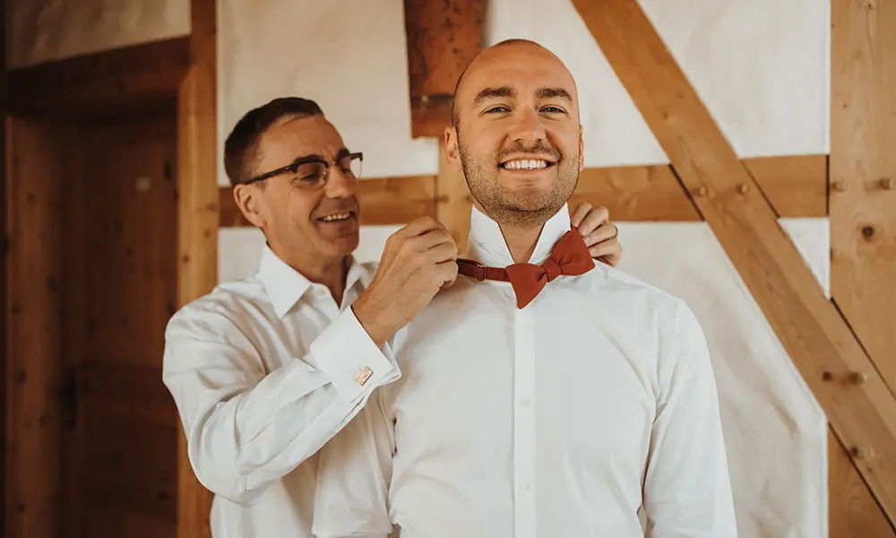 Ein Bräutigam lächelt glücklich, während ein älterer Herr ihm die rote Fliege bindet. Der rustikale Hintergrund mit Holzelementen sorgt für eine warme Atmosphäre.