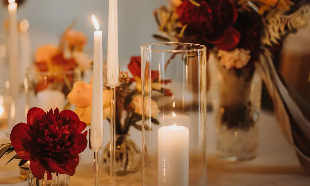 Elegante Hochzeitsdekoration: Eine Kombination aus roten und orangefarbenen Blumen in Vasen, begleitet von hohen Kerzen in Glaszylindern, schafft ein warmes, festliches Ambiente für die Tischgestaltung.