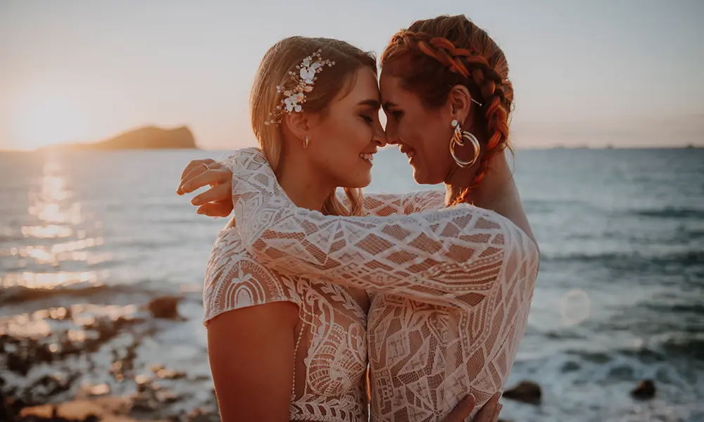 Romantischer Moment am Meer: Zwei Frauen in Brautkleidern halten sich lächelnd in den Armen vor der Kulisse eines Sonnenuntergangs. Die warmen Farben des Himmels und des Ozeans betonen die liebevolle Atmosphäre.