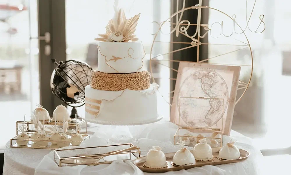 Reise-inspirierte Hochzeitstorte: Eine elegante, weiße Hochzeitstorte mit goldenen Akzenten, umgeben von Cake Pops und Desserts, dekoriert mit einer Globusfigur und einer Vintage-Weltkarte im Hintergrund.