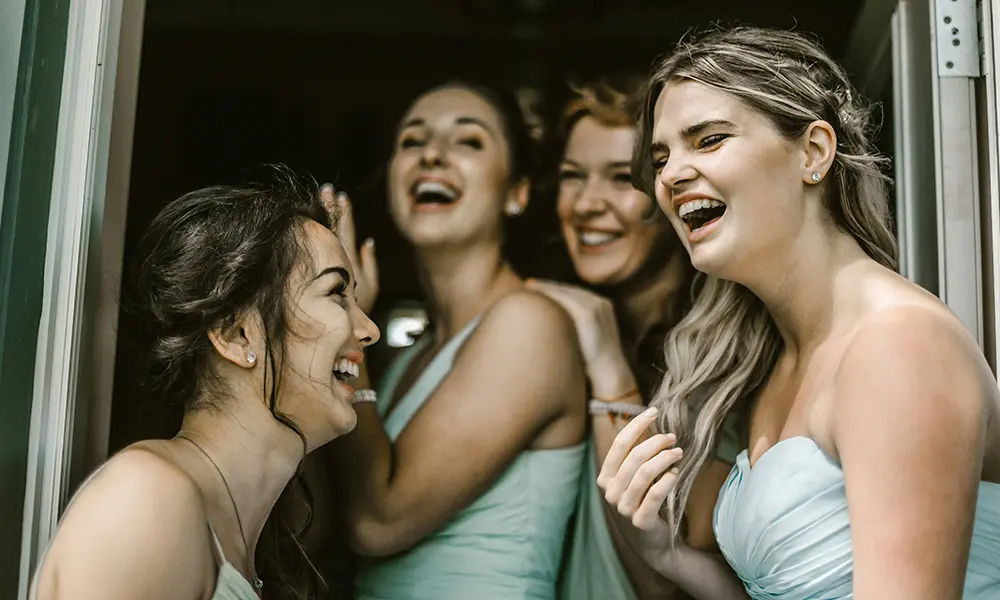 Lachende Brautjungfern: Eine Gruppe junger Frauen in hellblauen Kleidern scherzt und lacht zusammen, während sie entspannt in einer Tür stehen. Ein Moment voller Freude und Freundschaft.