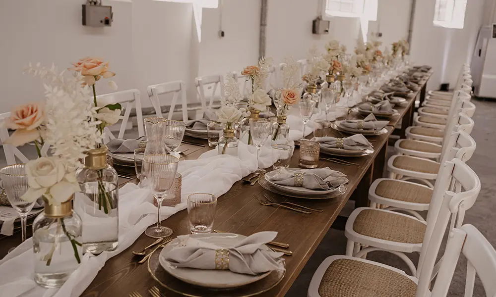 Lange Hochzeitstafel in modernem Stil: Dezente Dekoration aus weißen Rosen und Orangetönen, feines Geschirr, goldene Details und graue Servietten auf einer rustikalen Holzoberfläche.