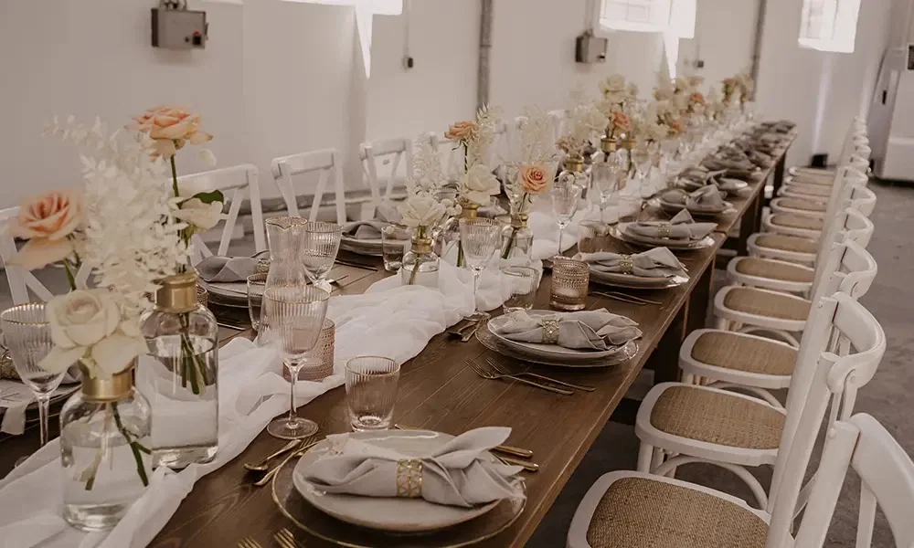 Lange Hochzeitstafel in modernem Stil: Dezente Dekoration aus weißen Rosen und Orangetönen, feines Geschirr, goldene Details und graue Servietten auf einer rustikalen Holzoberfläche.
