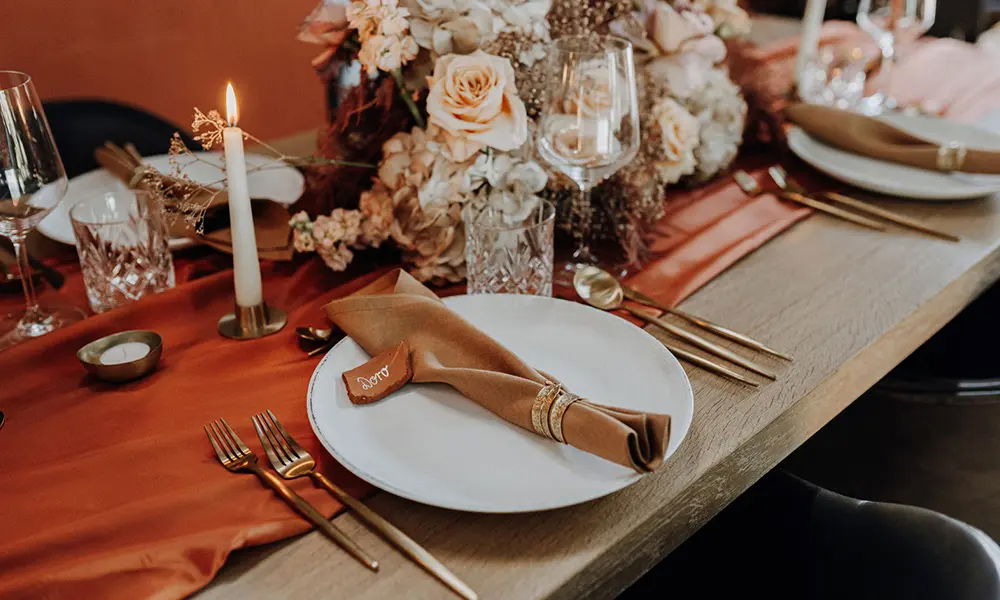 Elegante Hochzeitstafel mit herbstlichem Flair: Dekorativer Tischläufer in warmem Rot, goldfarbenes Besteck, eine stilvolle Kerze und ein opulentes Blumengesteck in dezenten Naturfarben zieren die Tafel.