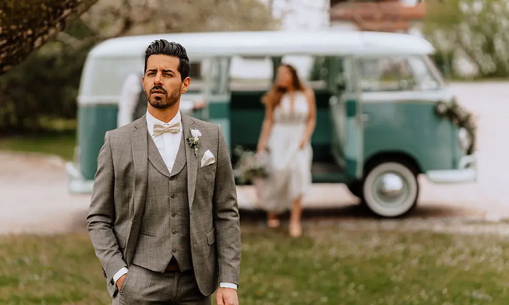 Hochzeitsfoto mit Retro-Charme: Der Bräutigam in einem eleganten grauen Anzug posiert vor einem dekorierten VW-Bulli, während die Braut im Hintergrund aussteigt und das Bild eine nostalgische Stimmung vermittelt.