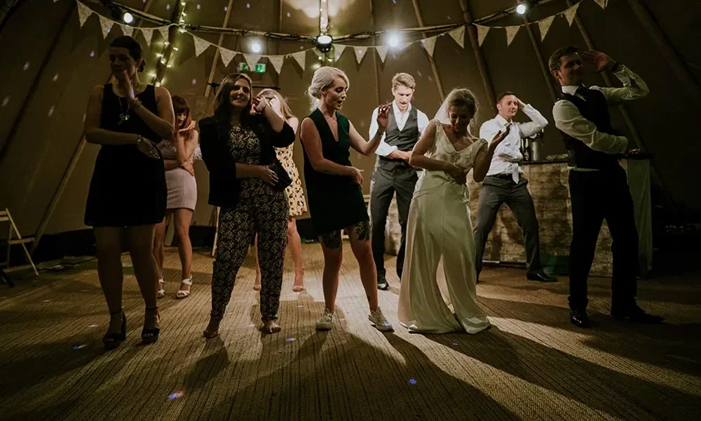Fröhliche Tanzszene bei einer Hochzeit: Die Braut und andere Gäste tanzen gemeinsam im Festzelt, das mit Lichterketten und Wimpelgirlanden dekoriert ist. Der Boden und die Beleuchtung schaffen eine gemütliche Stimmung.