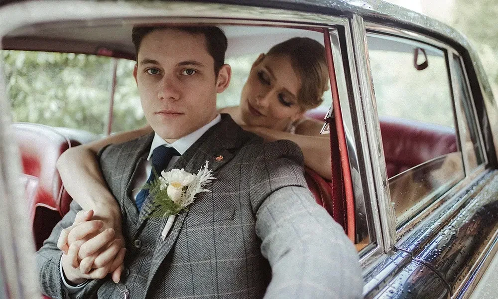 Eine romantische Hochzeitsaufnahme in einem Vintage-Auto: Der Bräutigam sitzt aufrecht mit einem nachdenklichen Blick, während die Braut ihn zärtlich umarmt. Die rote Lederausstattung des Fahrzeugs und die Regentropfen auf der Karosserie unterstreichen die besondere Stimmung des Moments.