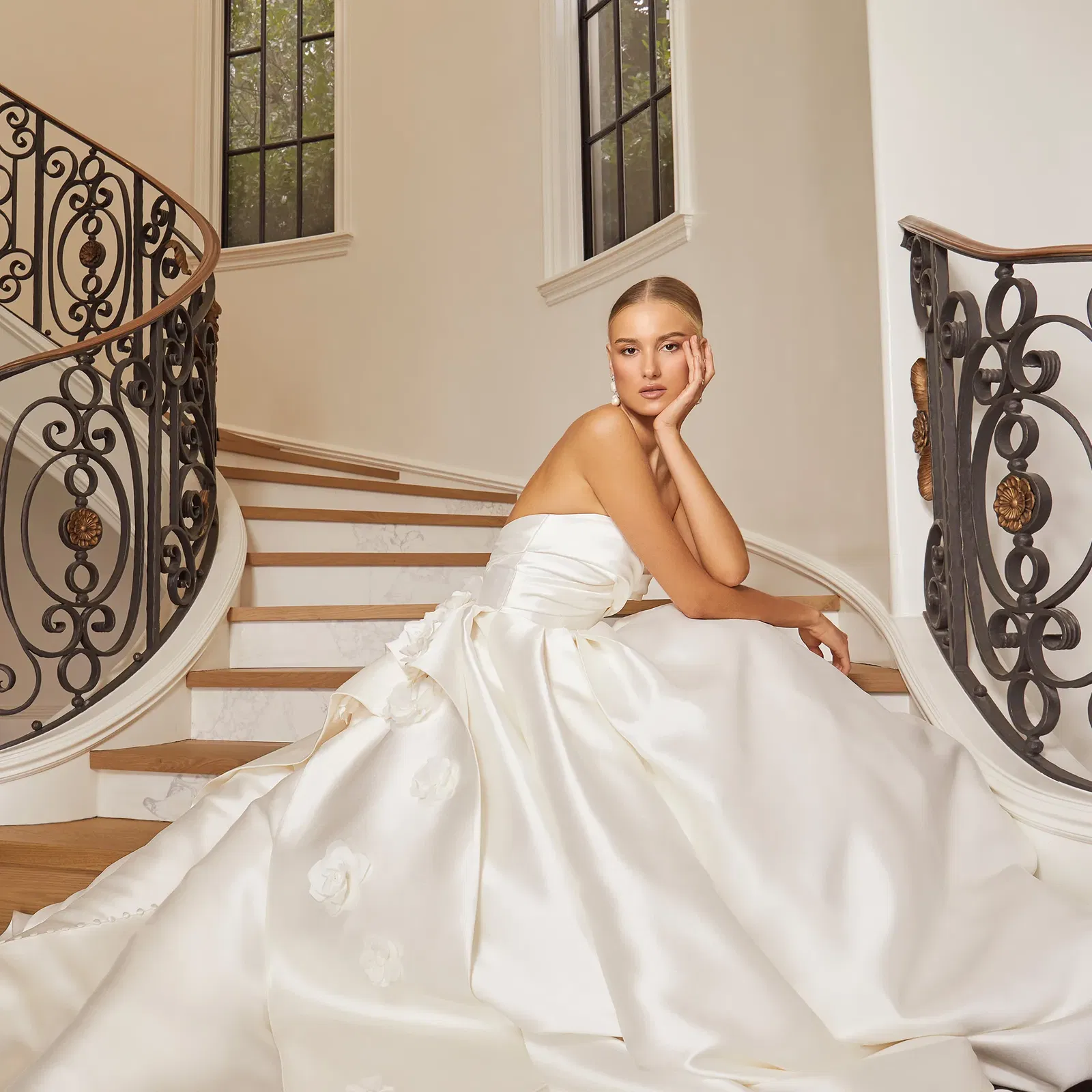 Die Braut sitzt auf der gleichen geschwungenen Treppe und blickt über die Schulter zurück. Sie trägt ein weites, fließendes Kleid mit einem Bandeau-Oberteil und einer auffälligen Schleppe. Das Kleid hat florale Applikationen, und die Braut trägt ein entspanntes Lächeln.