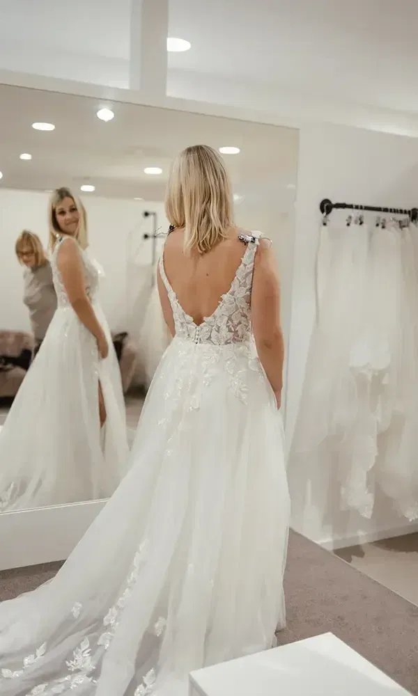 Lächelnde Braut bewundert ihr Hochzeitskleid im Spiegel, während eine Freundin den Anproben-Moment genießt.