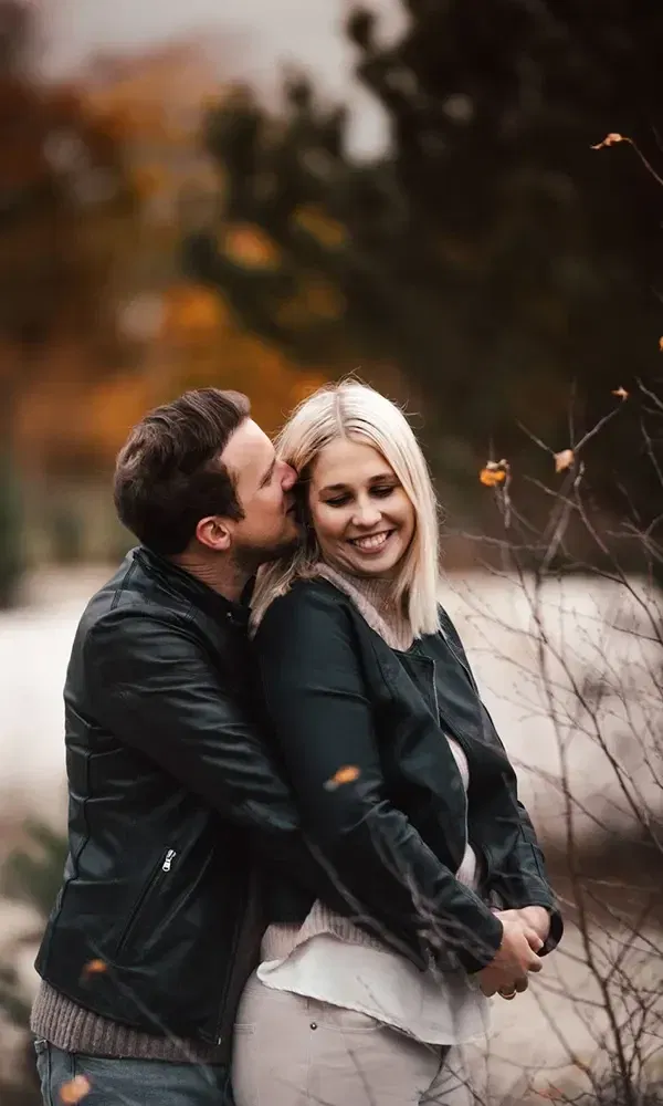 Liebevolles Bild eines Paares im Freien: Er küsst sie sanft auf die Wange, während sie glücklich lächelt.