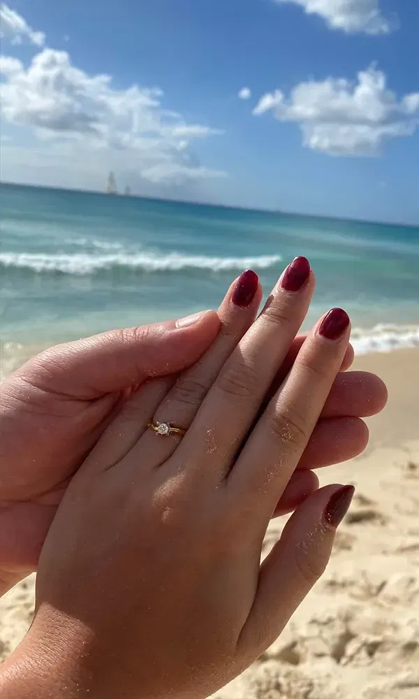 Eine Frau zeigt stolz ihren Verlobungsring, während sie von einer zweiten Hand gehalten wird, vor einer malerischen Küstenkulisse.