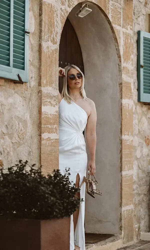 Junge Frau im stilvollen weißen Kleid, die entspannt in einem steinernen Torbogen lehnt, mit Sandalen in der Hand und Sonnenbrille auf.