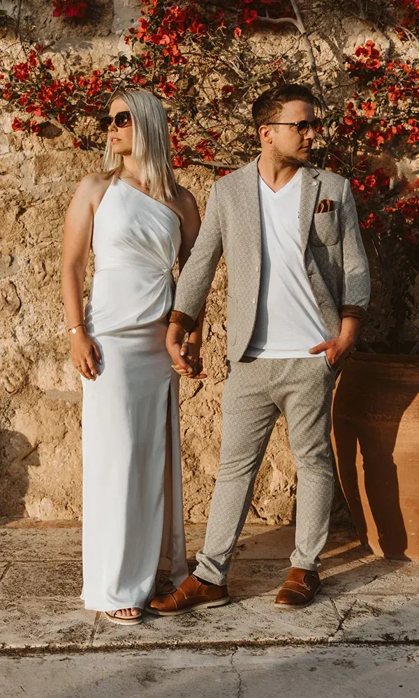 Ein verlobtes Paar posiert vor einer Wand aus Stein und roten Blumen. Die Frau hat ein elegantes weißes Kleid und Sonnenbrille, der Mann ist in einem grauen Anzug mit lässigem T-Shirt gekleidet.