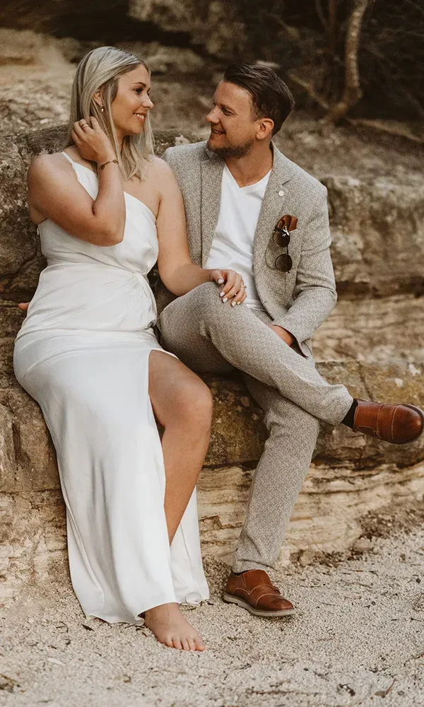 Paar in romantischer Szene: die Frau im weißen Kleid lehnt sich zum Mann im Anzug, beide strahlen sich auf einem Felsen an.