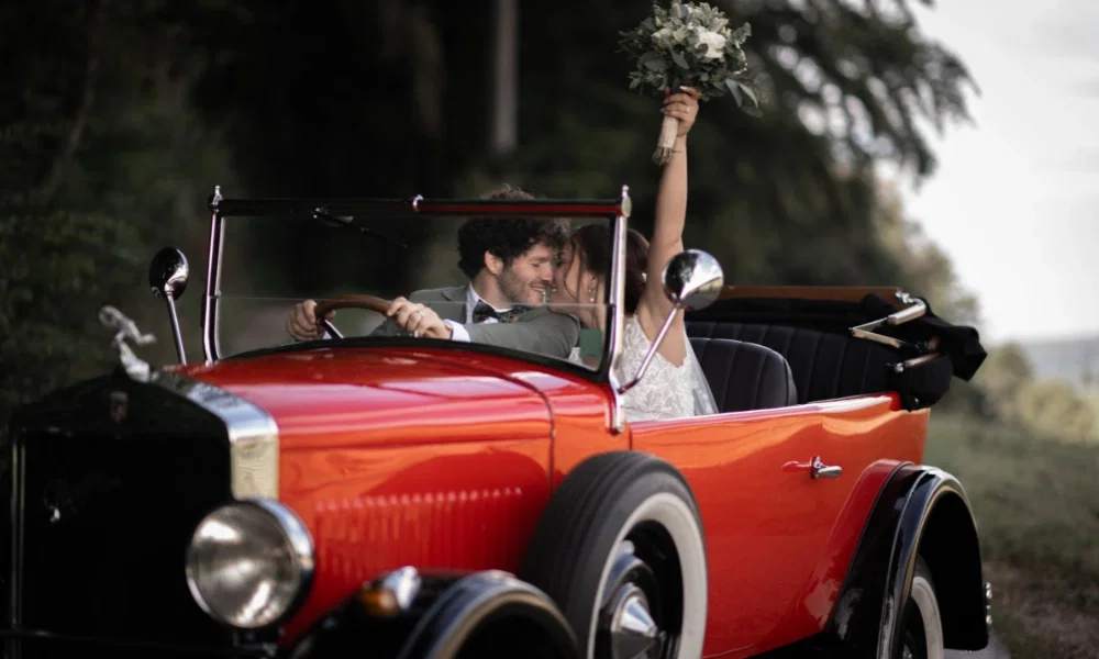 Das Brautpaar sitzt in einem roten Oldtimer. Sie schauen sich verliebt an und die Braut hält einen Blumenstrauss mit weissen Rosen in die Luft.