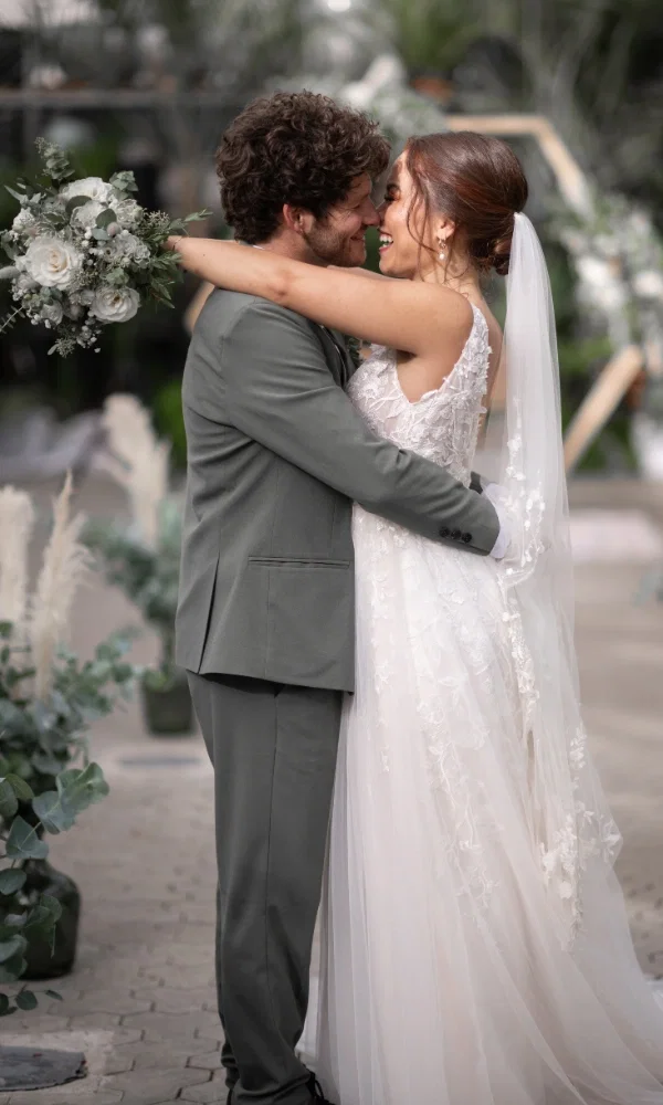 Ein Brautpaar umarmt sich glücklich bei ihrer Hochzeit. Der Braut trägt ein weisses Hochzeitskleid mit Spitzenverzierung und einen Schleier. Die Brut hält einen Blumenstrauss aus weissen Rosen.