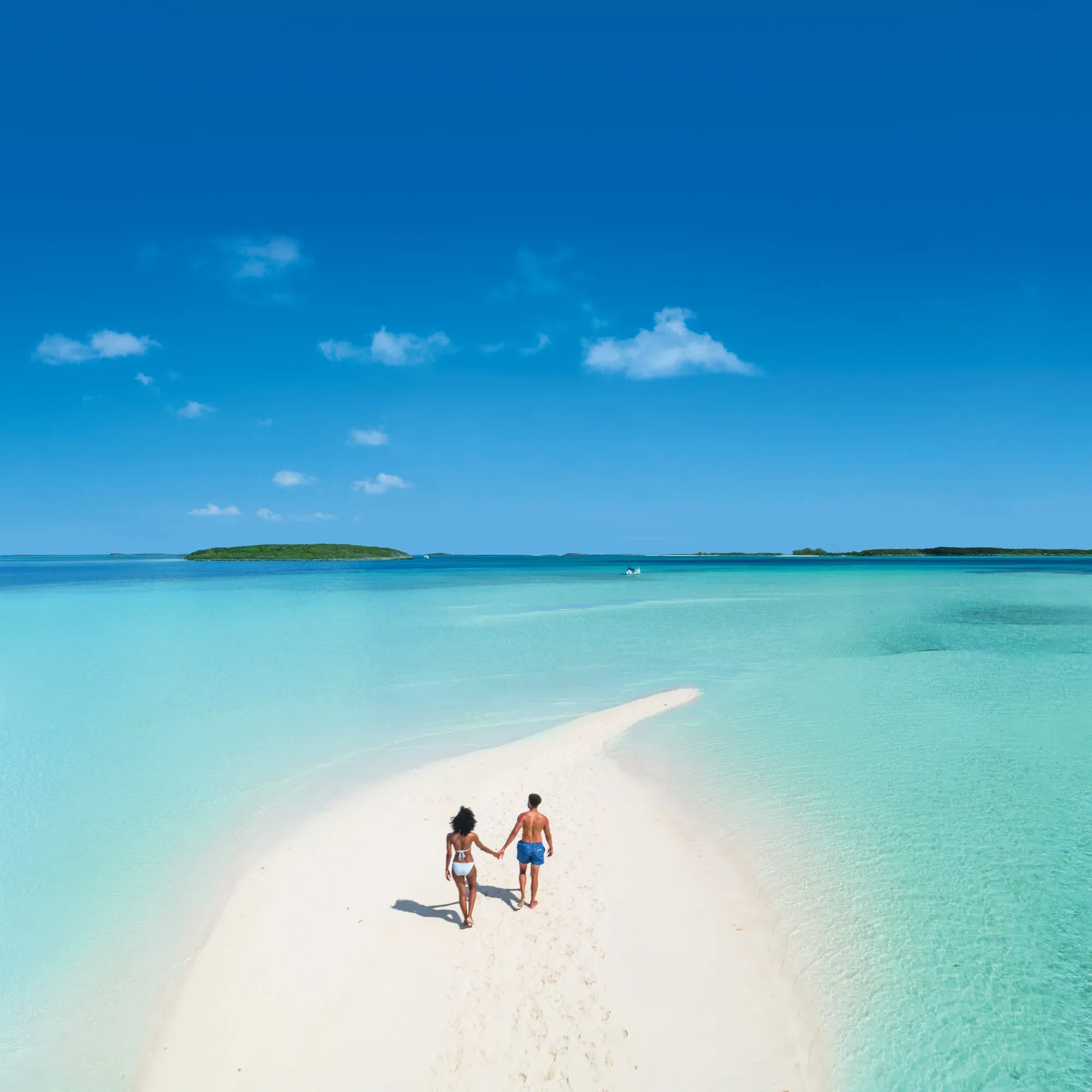 Mann und Frau in Badesachen laufen eine Sandzunge entlang. Drumherum türkises Meer.