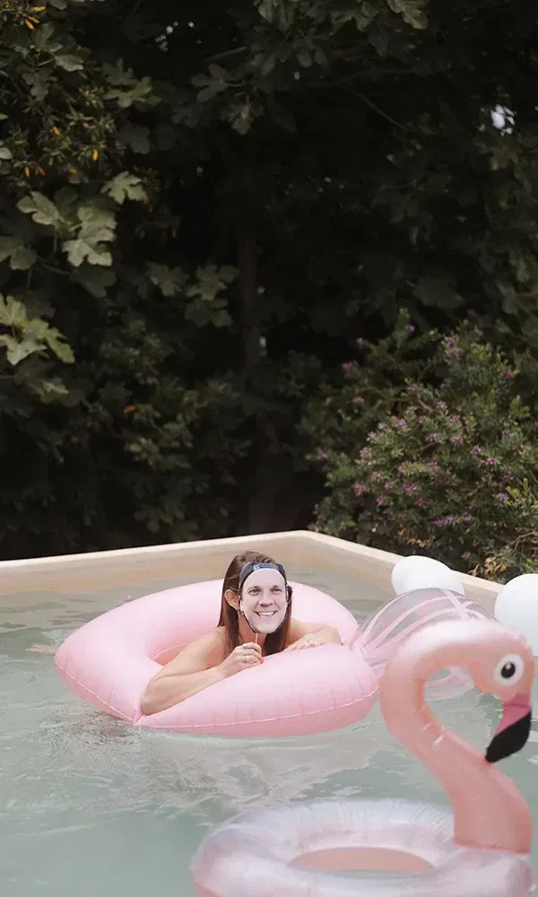Auf einem rosa Schwimmring im Pool liegt eine Person, die eine lustige Maske mit dem Gesicht eines Mannes trägt. Im Wasser ist ein aufblasbarer Flamingo zu sehen.