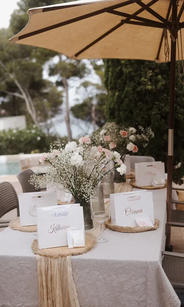 Tischdeko für eine Feier im Freien mit liebevoll gestalteten Gastgeschenken, Namenskarten und Blumenarrangements in Weiß und Rosa, umgeben von Natur.